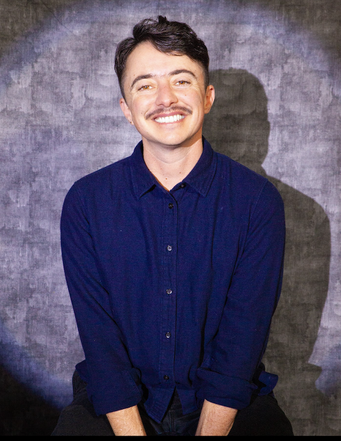 Photo of Dean Spade smiling in front of a grey background wearing a navy blue shirt.